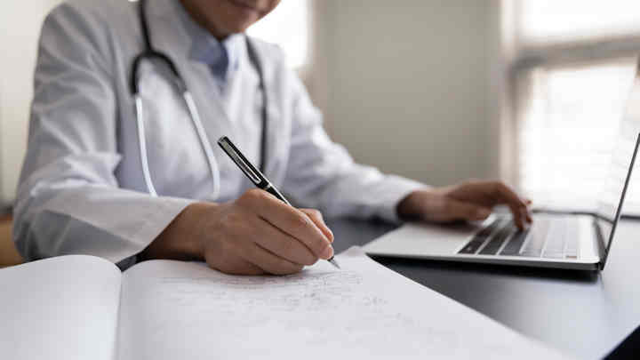 A doctor making notes from information on a laptop.