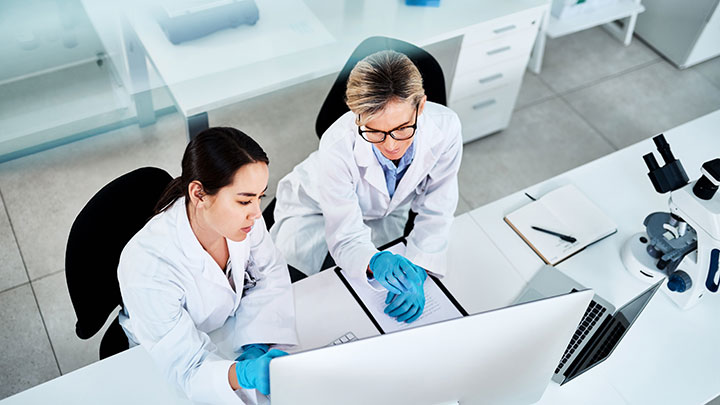 Two researchers in lab coats working together at a computer in a bright setting.