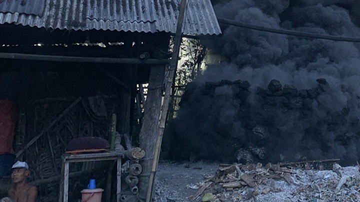 An Indonesian person sitting in a hut while smoke from a fire billows nearby.