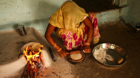 An Asian woman rolling out chapatis next to a cooking fire.
