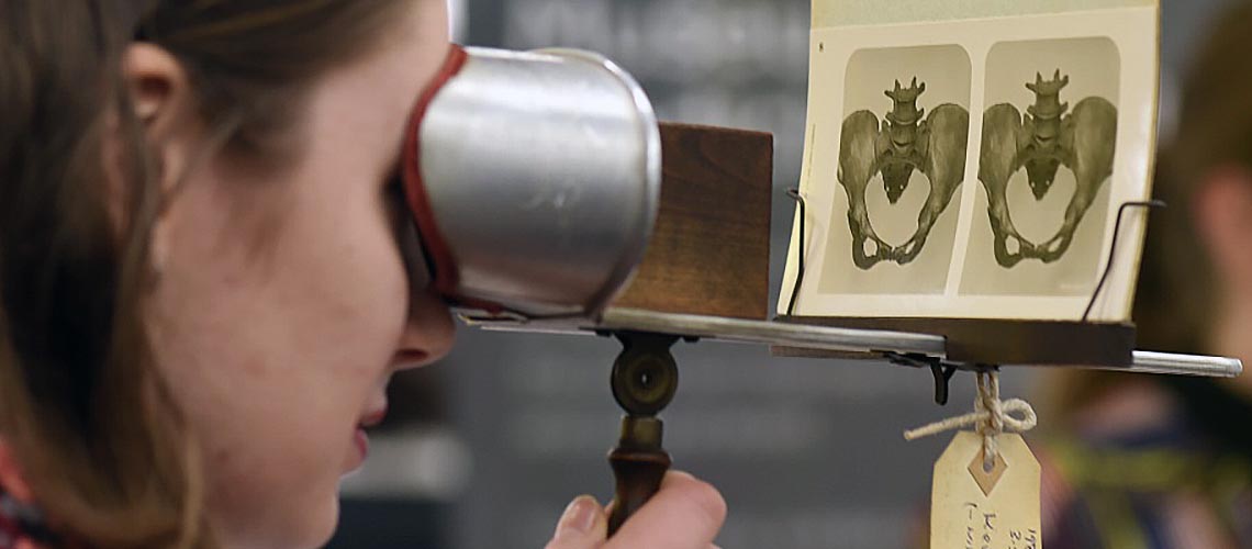 A young woman looks through a stereoscopic viewer, an anatomical teaching device. She examines a 3D image of the bone structure of an enlarged female pelvis, printed on a viewing card.