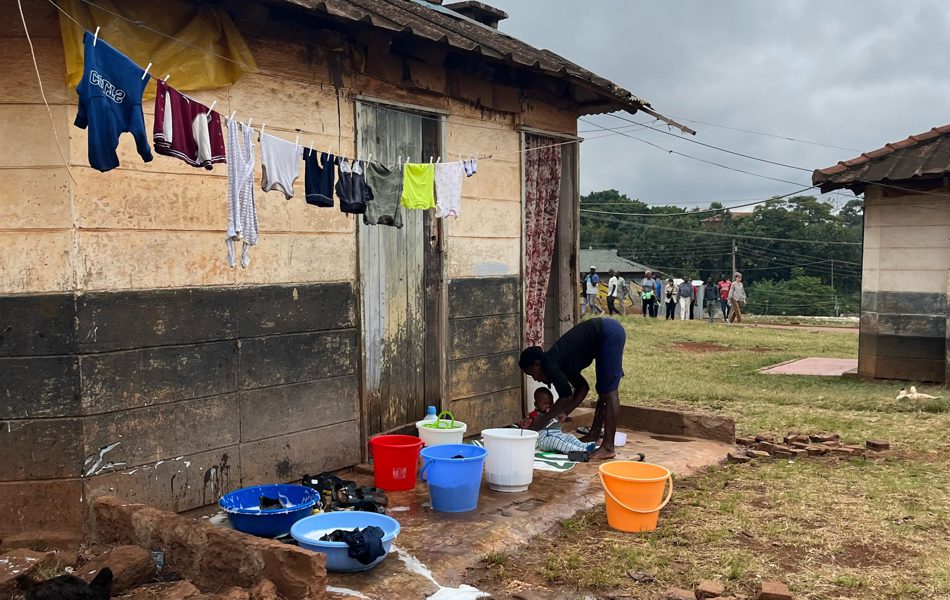 A woman with a child outside a house next to buckets of water for washing.