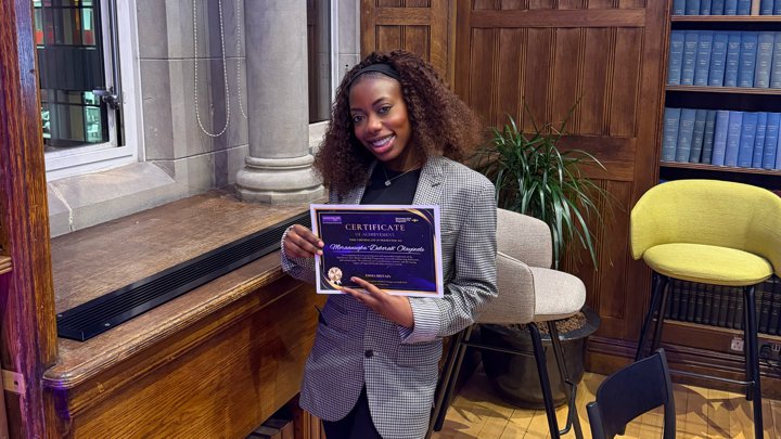 Tomi holding a certificate and smiling towards the camera in a wood-panelled room.