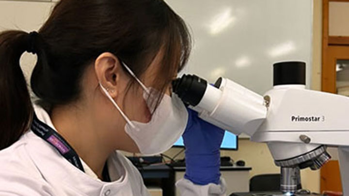 Balqis looking through a microscope whilst wearing a white face mask.