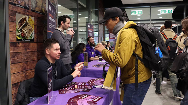 A man in a yellow jacket and black cap talking to another man sitting at a stall at an open day.