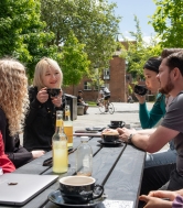 A group of researchers sat at a bench outside in the sun, laughing and smiling while enjoying coffees and drinks.