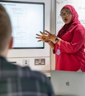 A female postgraduate researcher wearing red presents in front of a screen as someone in the foreground watches on