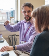 A male postgraduate researcher sat at a computer looks at their supervisor while discussing something, using their hand to illustrate the point they are making