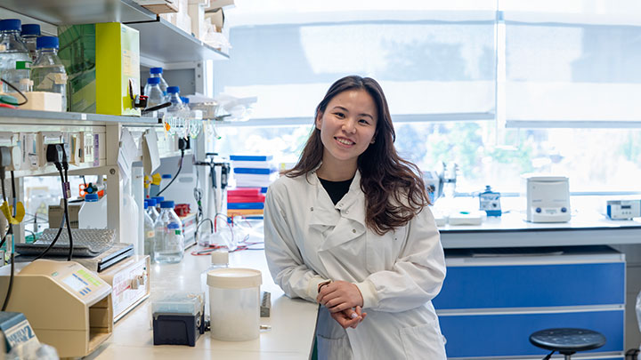 A postgraduate researcher leaning on a counter in a lab, smiling towards the camera.