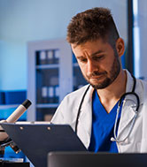 A male doctor with a beard looking down at a clipboard in a lab setting.
