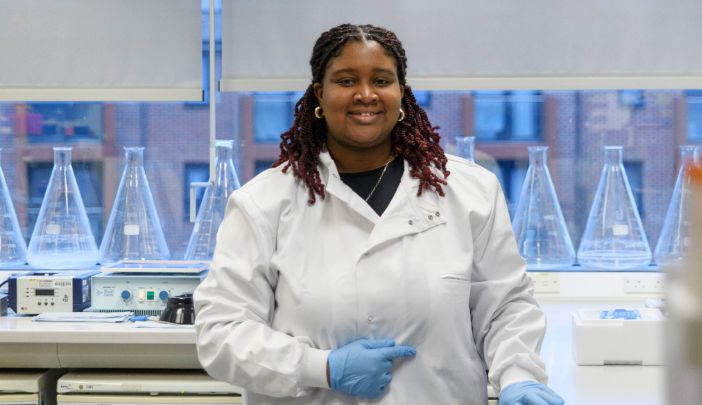 A researcher stood in a lab wearing a white lab coat smiling at the camera