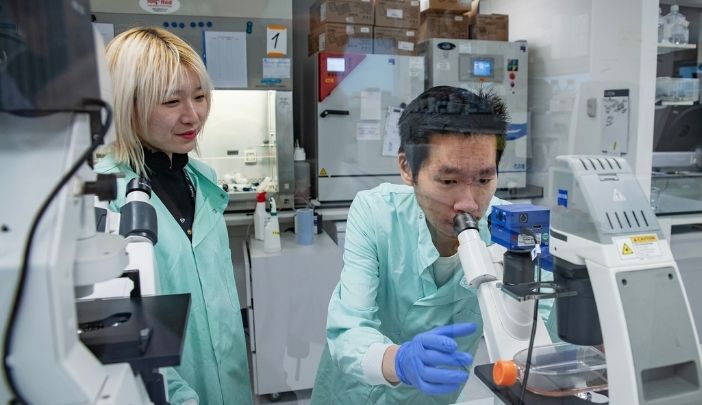 Two researchers in lab coats, one looking into a microscope, the other observing