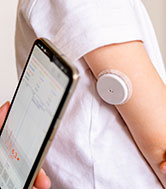  A person's arm with a white blood sugar device attached, with a smartphone being held on the left.