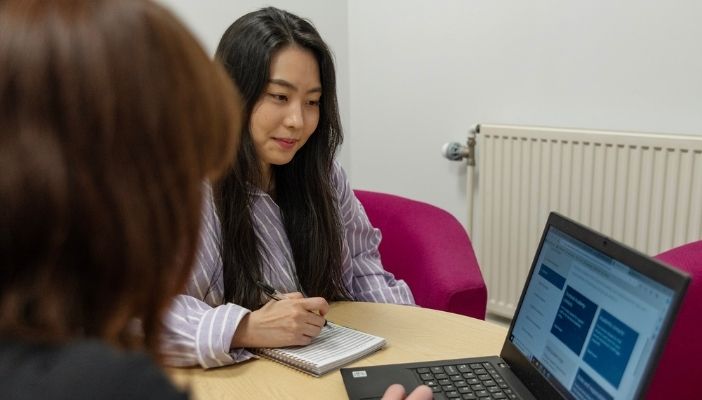 A member of the Doctoral Academy team sits at their laptop while a PGR writes on a notepad.